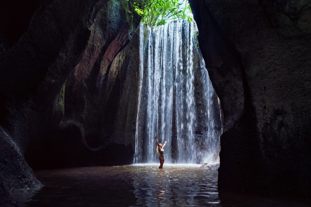 woman stand in underground cave pool under falling fresh water of tukad cepung waterfall. nature day tour, hiking activity adventure and fun at family tourist camp on summer vacation in bali island