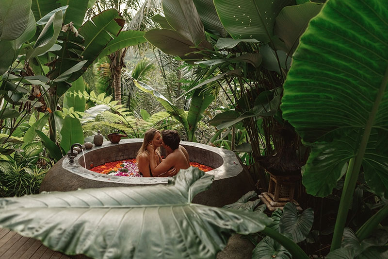 couple kissing in a tropical spa bath with flower pedals surrounded by tropical green trees and plants. 